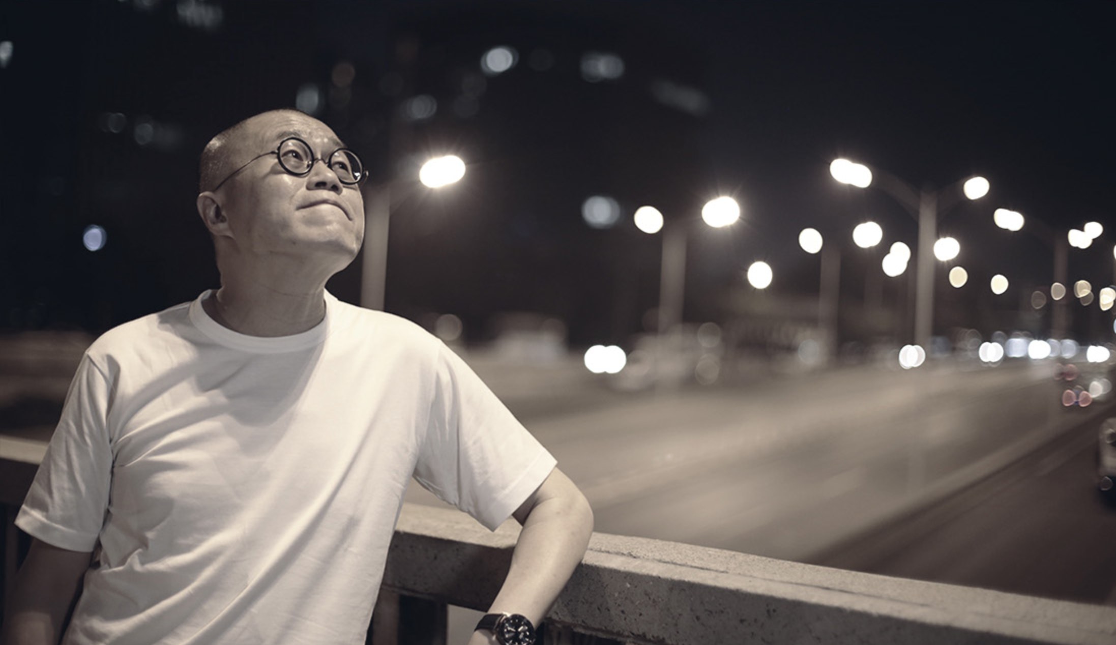 梁文道在夜晚街头阅读，身后有疾驰车辆和路灯
